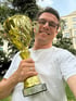 Ein lächelnder Mann mit Brille hält einen goldenen Pokal mit der Aufschrift "Cyber Academy 2023 Champion Cup" in einem Park.