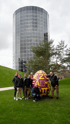 Eine Gruppe von acht Männern posiert neben einem großen, rot-gelben Osterei vor einem gläsernen Hochhaus.