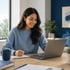 Smiling woman in a blue sweater taking notes in a notebook while looking at a laptop computer on a wooden desk.