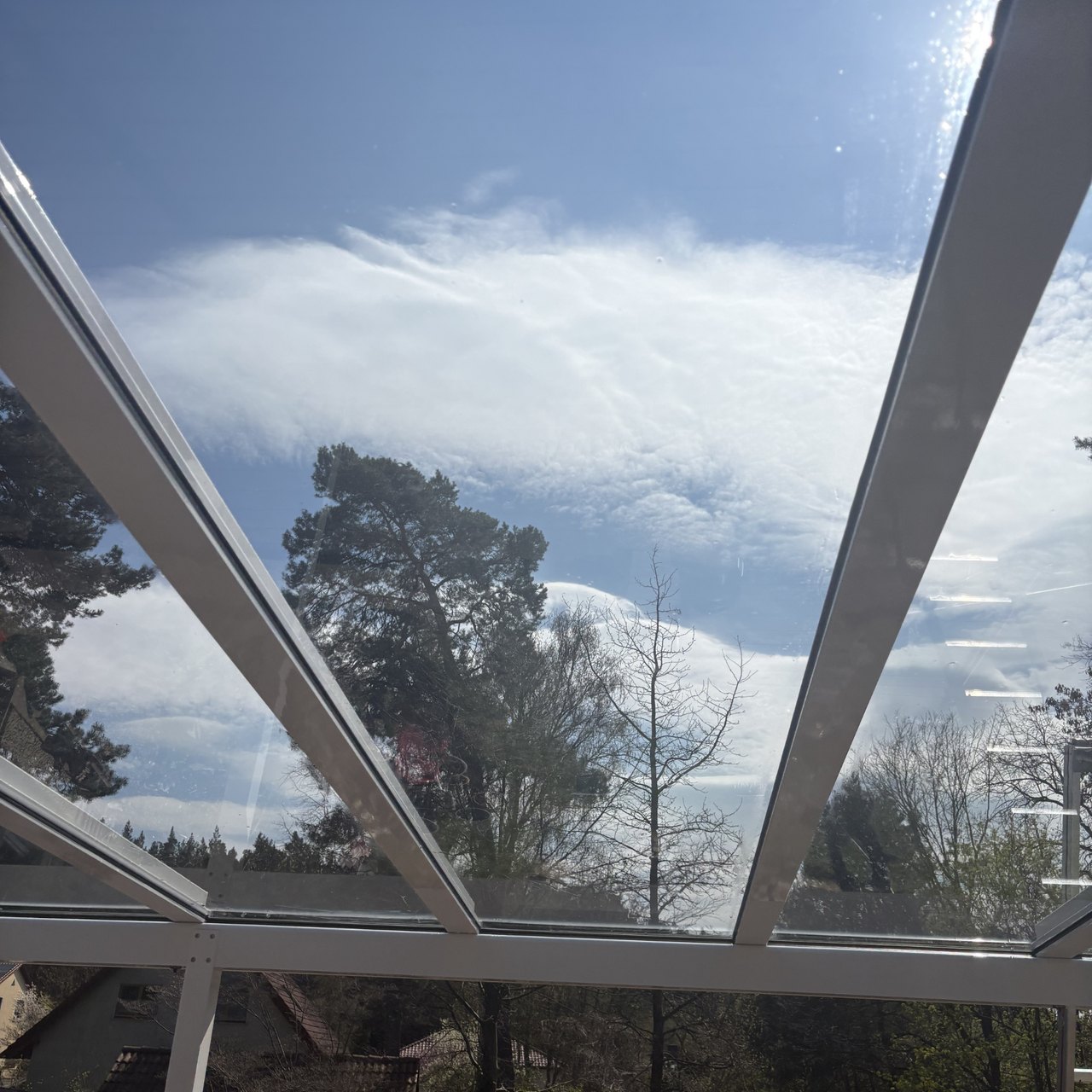 Blick durch ein Glasdach auf einen blauen Himmel mit weißen Wolken und grünen Bäumen.