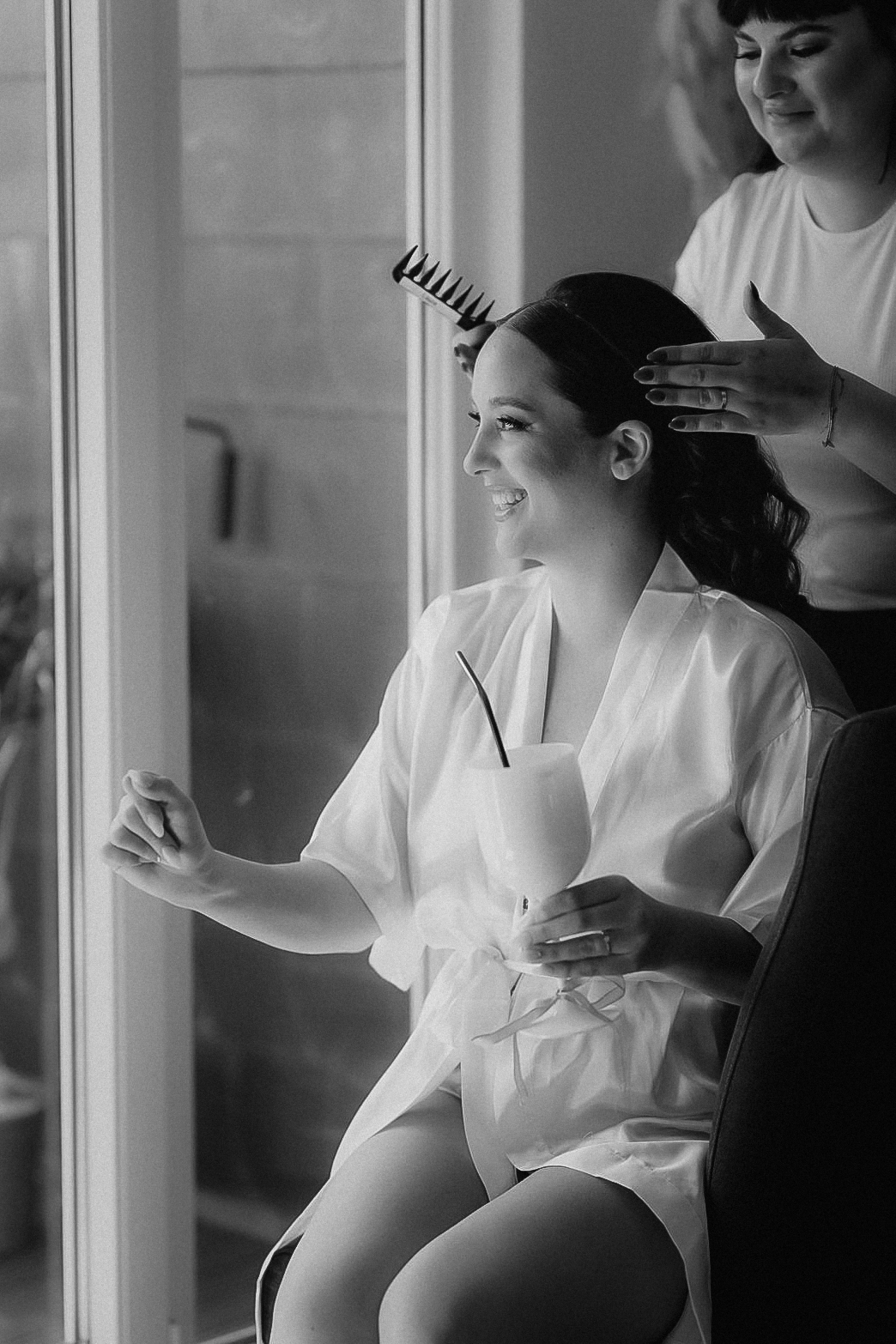 Schwarz-Weiß-Aufnahme: Die Braut wird am Fenster frisiert und lacht, im Satin-Morgenmantel und mit dem „Bride“-Glas in der Hand, beim Getting-ready in Köln.