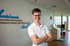 Lächelnder Mann mit Brille und weißem Poloshirt vor einer Wand mit blauem Schriftzug in einer modernen Praxis.