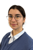 Eine junge Frau mit Brille und dunklem Haar, die ein blaues Hemd und einen blauen Pullover trägt.