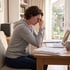 A woman sits at a desk with a laptop and stacks of paper, holding the bridge of her nose with her eyes closed.