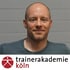 Glatzköpfiger Mann in grauem T-Shirt, lächelnd vor einer grauen Wand, mit "trainerakademie köln"-Logo darunter.