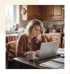 A woman with a furrowed brow works on a laptop at a kitchen table with a steaming mug and notebooks.