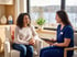 A female healthcare professional in blue scrubs speaks with a woman in a seated consultation by a window.