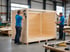 Zwei Handwerker in blauen T-Shirts montieren eine große Transportkiste aus Holz in einer Industriehalle.