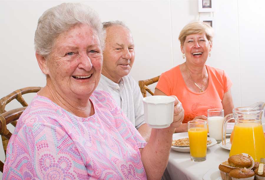 Drei ältere Menschen sitzen gemeinsam mit Kaffee und Saft an einem gedeckten Tisch.