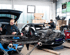 Zwei Mechaniker arbeiten in einer Werkstatt an einem schwarzen Auto; der Kofferraum ist offen, Teile liegen davor.