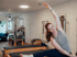 Eine Frau dehnt sich lächelnd auf einem Pilates-Reformer in einem hellen Studio mit Trainingsgeräten.