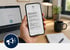 Hand hält Smartphone mit einer Textnachricht zu einem Krisenstabs-Meeting auf einem Schreibtisch mit Laptop - Software für Alarmierung - Notfall- und Krisenmanager (NKM®).