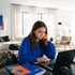 A girl in a blue sweatshirt looks at a laptop screen with a sad expression while sitting at a desk with notebooks.