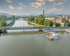 Fluss mit hölzernem Steg, grünen Ufern, Booten am Anleger und Industriegebäuden im Hintergrund unter bewölktem Himmel.