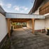 Ein moderner Carport aus Holz mit Flachdach und grauer Metallblende steht zwischen zwei Hauswänden auf gepflastertem Boden.