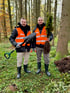 Zwei Männer in Warnwesten und Stiefeln stehen in einem Wald, einer hält eine Schaufel, der andere einen kleinen Baum.