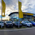 Opel Autohaus mit parkenden Neuwagen, gelben Opel-Flaggen und "OPEL" Schriftzug an der Fassade.