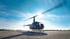 Ein Hubschrauber steht mit rotierenden Rotorblättern bei Gegenlicht auf einem Flugplatz unter blauem Himmel.