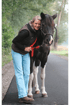 Eine lächelnde Frau mit grauen Haaren, brauner Weste und Jeans umarmt ein dunkles Pferd mit Halfter auf einem Weg.