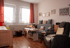 Wohnzimmer mit grauem Sofa, Sessel, Holztisch und zwei Fenstern mit roten Vorhängen vor einer hellen Wand mit gerahmten Fotos.