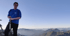 Mann in blauem Sweatshirt und dunkler Hose steht auf einem Berggipfel und hält Wanderstöcke.