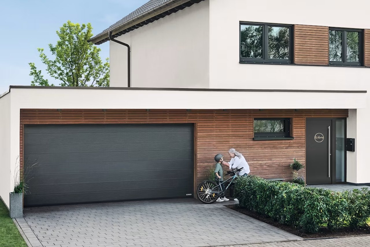 Anthrazitfarbenes Hörmann Sektionaltor an einem Wohnhaus mit Holzfassade, Mutter und Kind mit Fahrrad in der Einfahrt