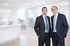 Zwei Männer in dunklen Anzügen und Krawatten lächeln in einem hellen, unscharfen Büro.