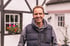 Ein lächelnder Mann in einer dunklen Weste und einem Kapuzenpullover steht vor einem weißen Fachwerkhaus.