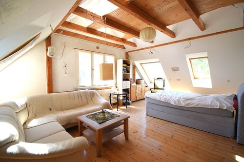 Dachboden-Wohnzimmer mit hellen Wänden, Holzbalken, zwei Sofas, einem Holztisch und einem Bett unter dem Dachfenster.