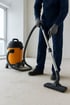 Eine Person in Arbeitskleidung saugt mit einem orangefarbenen Nass-Trocken-Sauger einen staubigen Boden.