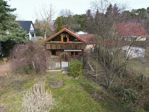 Kleines braunes Holzhaus mit orangem Dach und Balkon, umgeben von trockenen Bäumen und grünem Gras.
