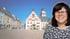 Lächelnde Frau mit Brille vor einem Marktplatz mit Rathaus und barocken Gebäuden.