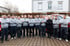 Eine Gruppe von 16 Mitarbeitern in grauen Uniformen steht mit verschränkten Armen vor roten Lieferwagen und einem Gebäude.