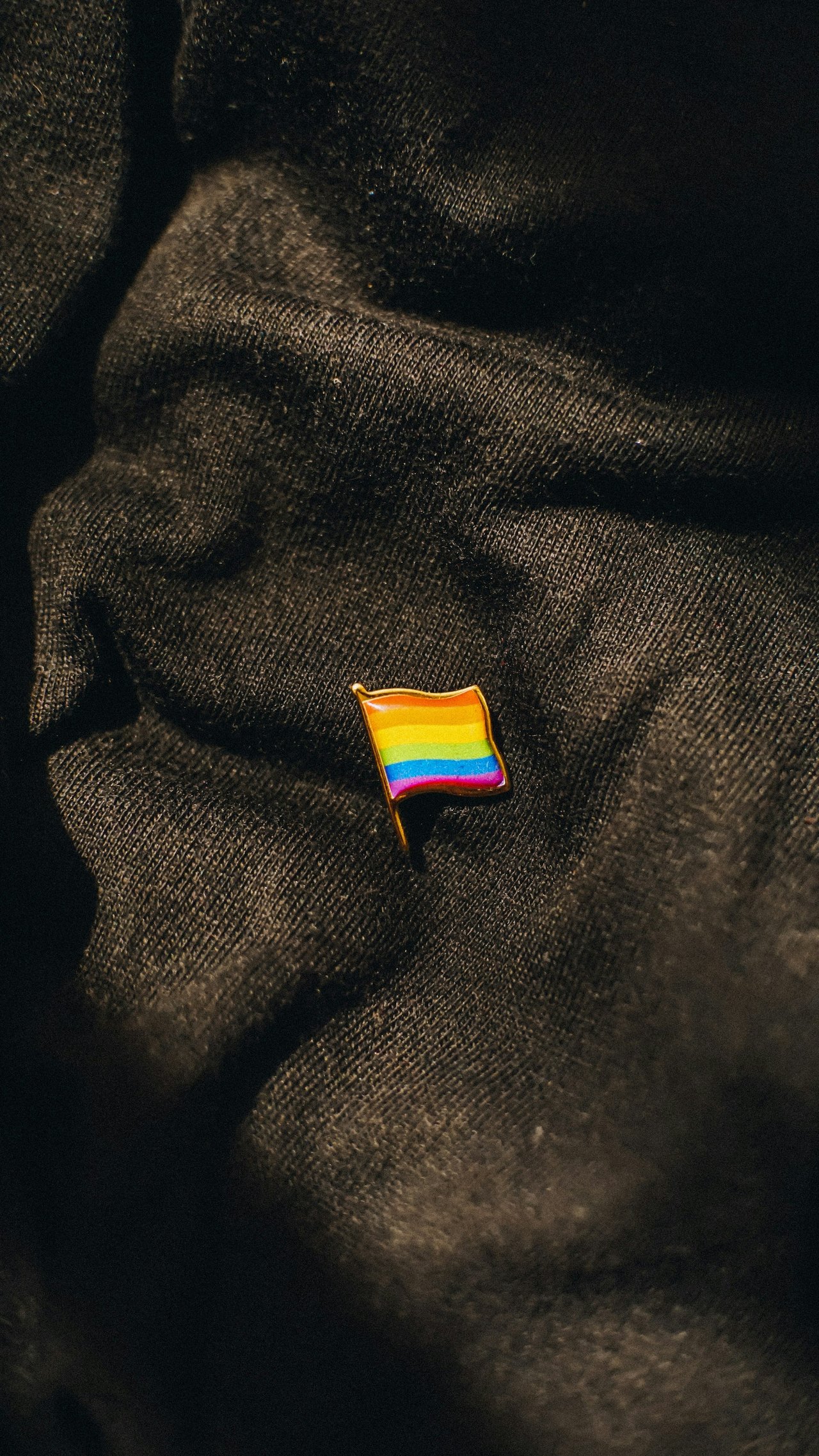 A rainbow flag pin, also known as a pride flag, on crumpled black fabric.