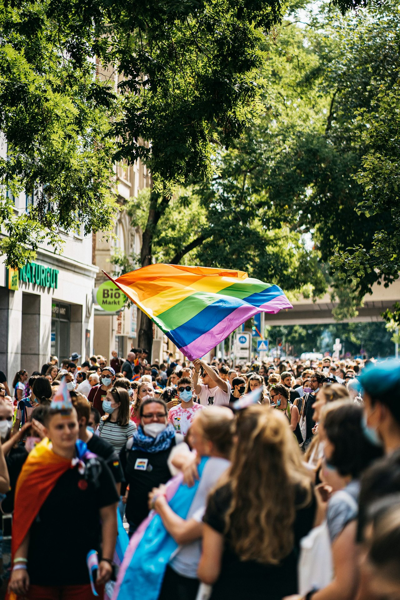 Viele Menschen auf der Straße, eine Person hält eine große Regenbogenflagge über der Menge.