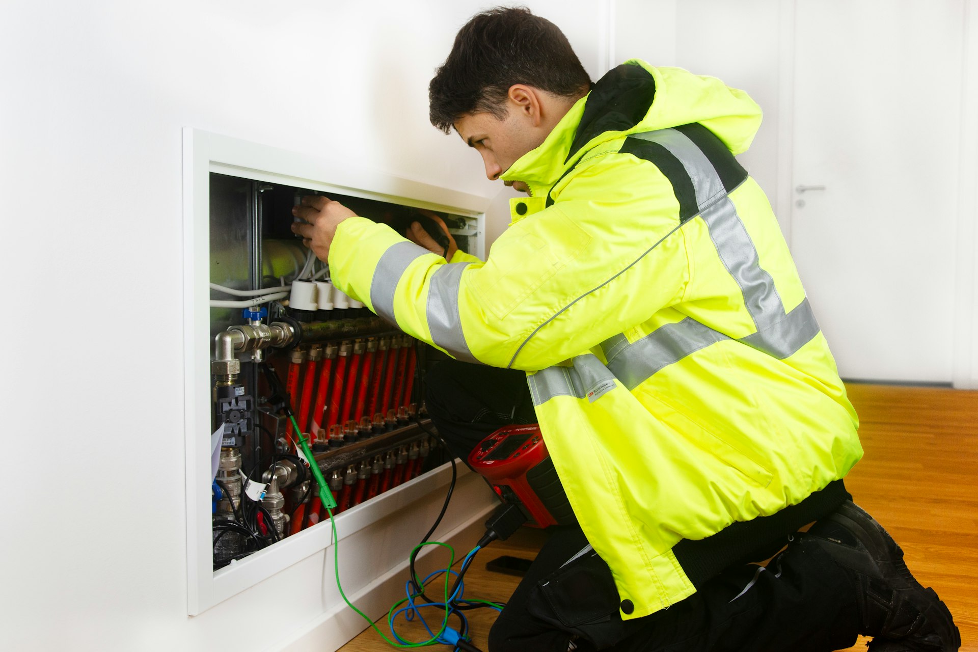Monteur in Warnjacke arbeitet kniend an einem geöffneten Heizkreisverteiler in einer Wohnung, am Boden ein Messgerät mit Prüfleitungen